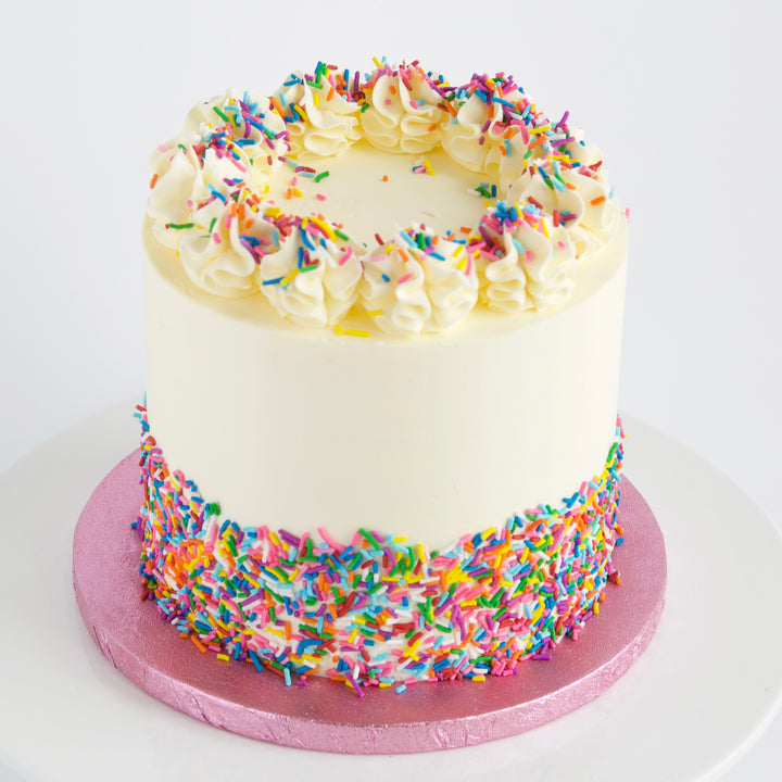 Close up of Vanilla Rainbow Cake, covered in Swiss meringue buttercream and rainbow sprinkle. Finished with a circle of buttercream rosettes