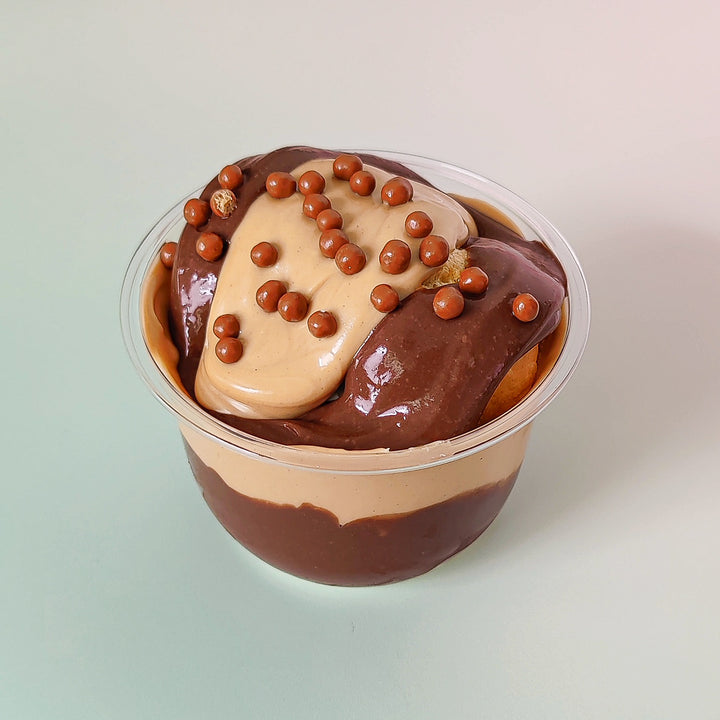 Hazelnut and chocolate profiteroles in bioplastic pots, available for delivery across London. Fresh choux bun pastry filled with vanilla creme patissiere and covered with chocolate sauce and our white hazelnut cream, with crispy milk chocolate pearls. Order online at Punk Cake for dessert delivery.